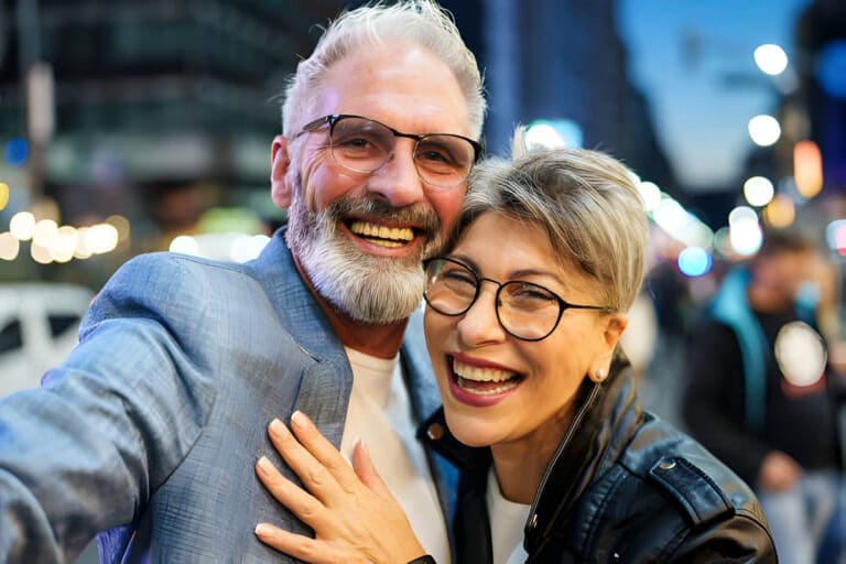 A couple both wearing glasses smile at the camera as they have their picture taken in the city.