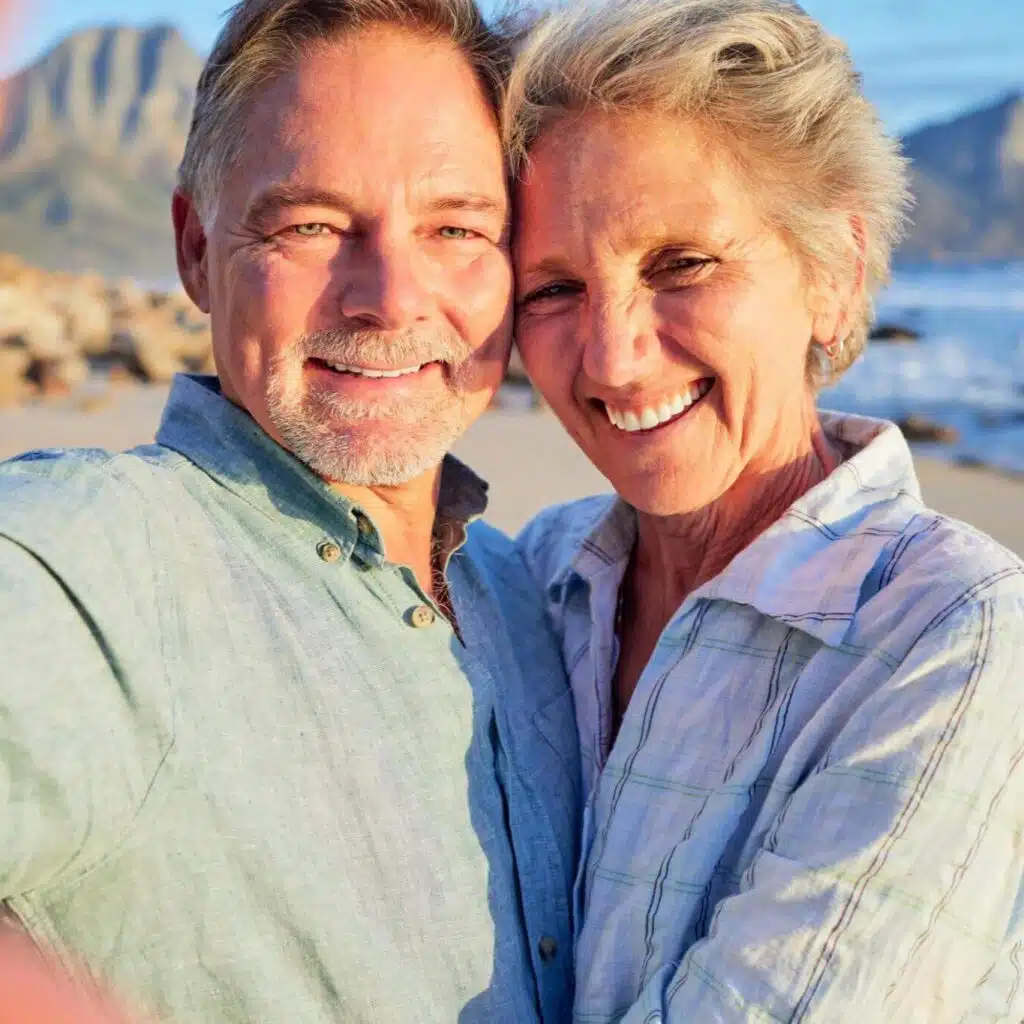 A happy couple takes a selfie on the beach as they smile in the morning sun.