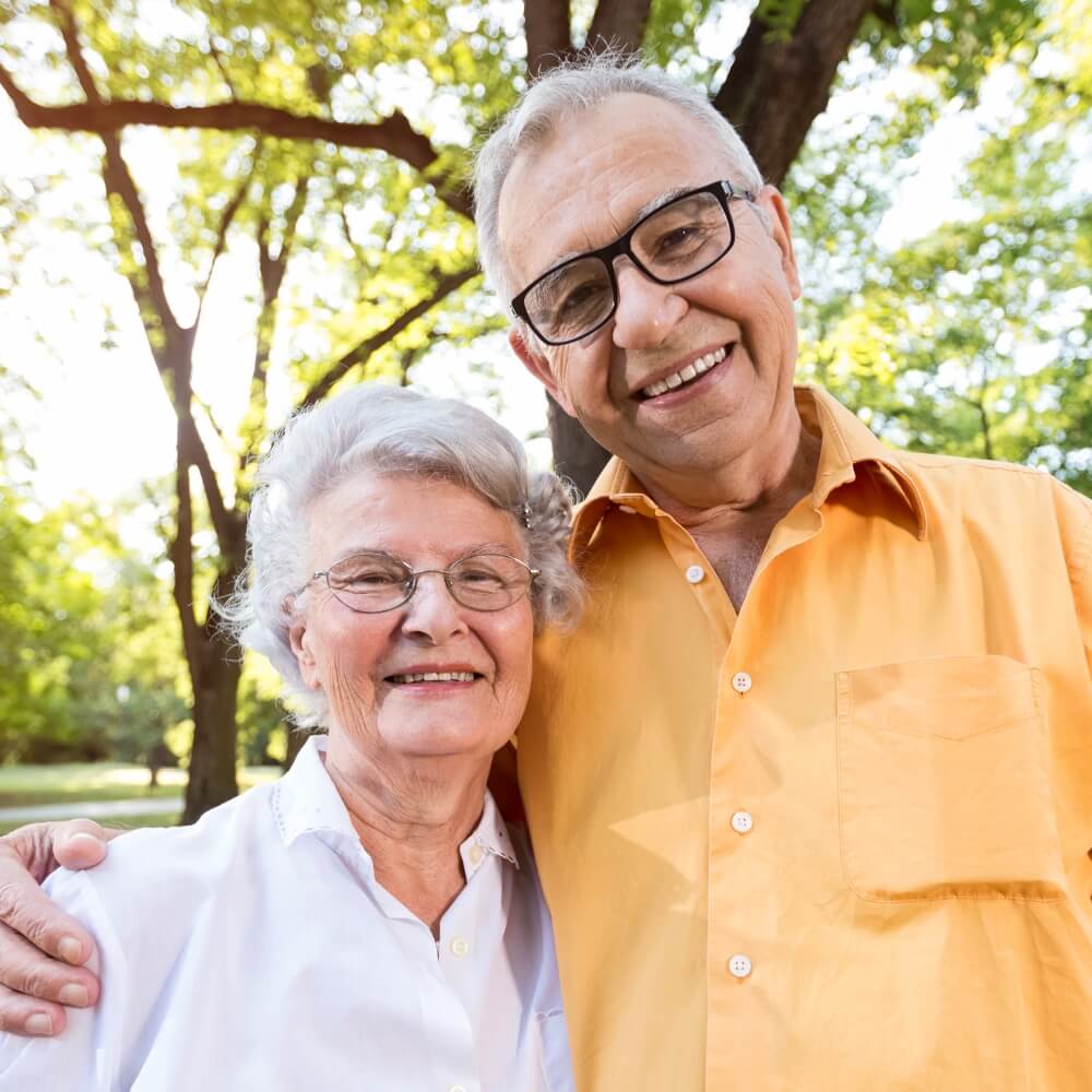 A elderly couple stands in front of a tree smiling, one wearing white and the other orange.