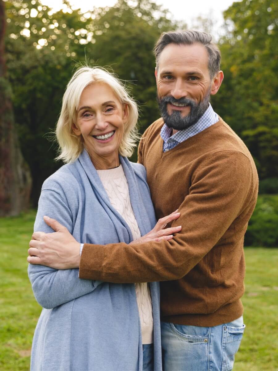 A middle-age couple hug on the lawn while smiling at the camera.