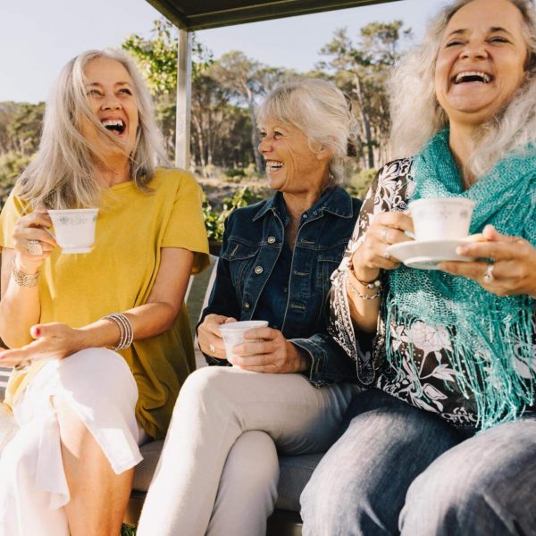 Three female friends drink tea and laugh at jokes on a cool but sunny afternoon.